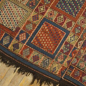 Intricate flatweave rug with repeating diamond and square patterns in red, blue, and gold on light hardwood.