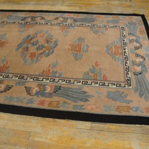 Tan field rug with blue and coral stylized motifs and a wide black border resting on yellow wood planks.