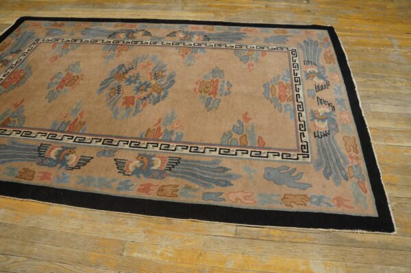 Tan field rug with blue and coral stylized motifs and a wide black border resting on yellow wood planks.