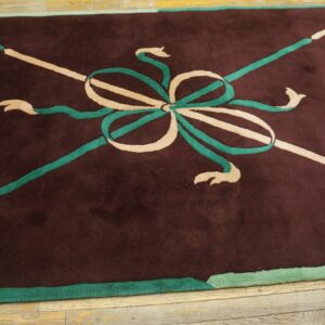 Low-pile maroon rug with a stylized beige and green central bow design resting on wood planks.
