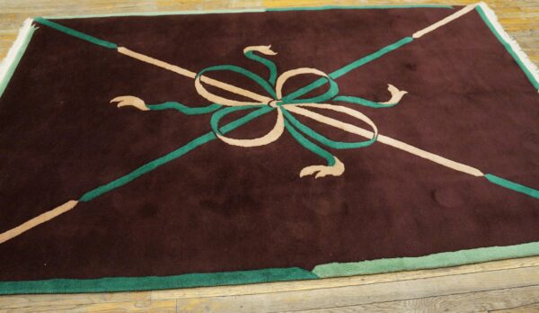 Low-pile maroon rug with a stylized beige and green central bow design resting on wood planks.