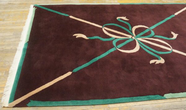 Maroon rug featuring a stylized tan and teal bow pattern and white fringe on worn wood flooring.