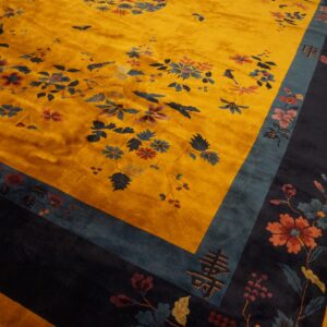 Gold rug with high pile and dark blue floral motifs, bordered by teal and navy on wood planks.