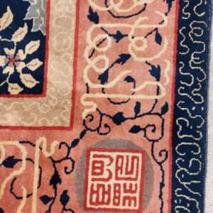 Corner detail of a patterned rug featuring salmon pink, navy blue, and tan borders and a seal motif.