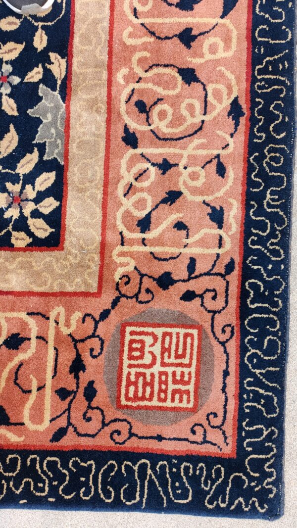 Corner detail of a patterned rug featuring salmon pink, navy blue, and tan borders and a seal motif.