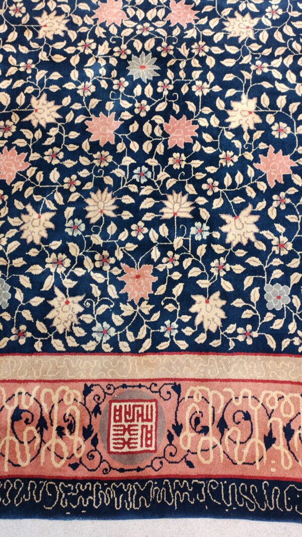 Dark blue rug with dense floral pattern and a dusty pink border featuring a symbolic cartouche.