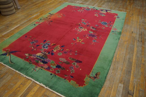 Red rug with green border and multicolor botanical motifs, including birds, sits on wood flooring.