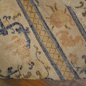 Beige low pile rug featuring dark blue and orange stylized motifs, a striped border, and visible fringes on a wood floor.