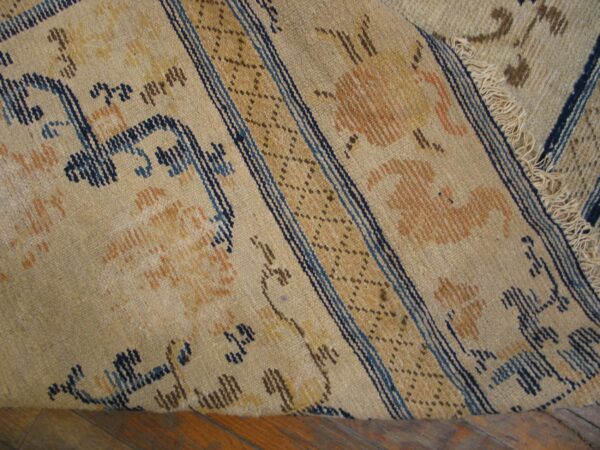 Beige low pile rug featuring dark blue and orange stylized motifs, a striped border, and visible fringes on a wood floor.