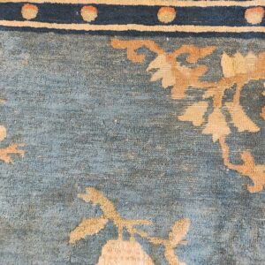 Close detail of a blue, low-pile rug with tan floral motifs and a border strip of navy, cream, and peach.