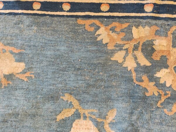 Close detail of a blue, low-pile rug with tan floral motifs and a border strip of navy, cream, and peach.