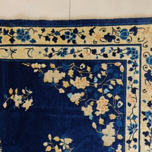 The corner of a deep blue floral rug with a contrasting cream and blue botanical border on light tile.