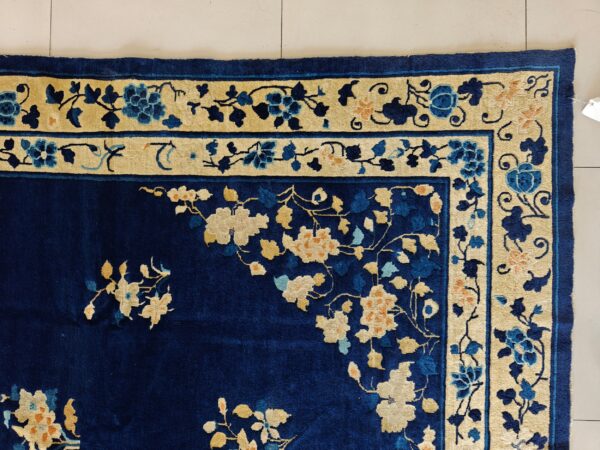 The corner of a deep blue floral rug with a contrasting cream and blue botanical border on light tile.