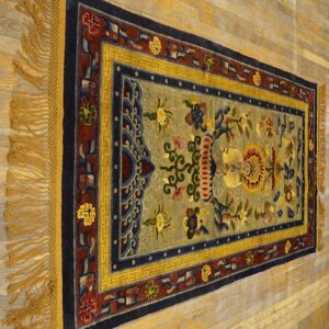 Muted sage rug with navy, red, and gold ornamental borders and fringe on light hardwood flooring.
