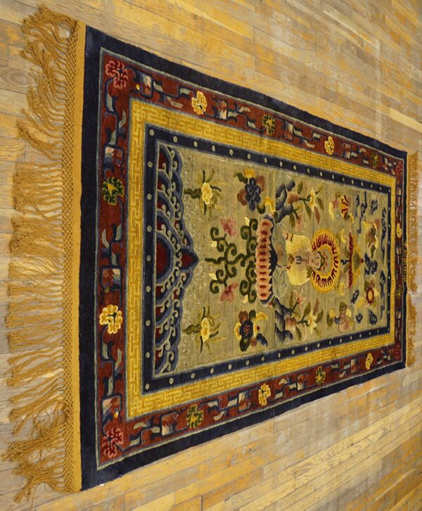 Muted sage rug with navy, red, and gold ornamental borders and fringe on light hardwood flooring.