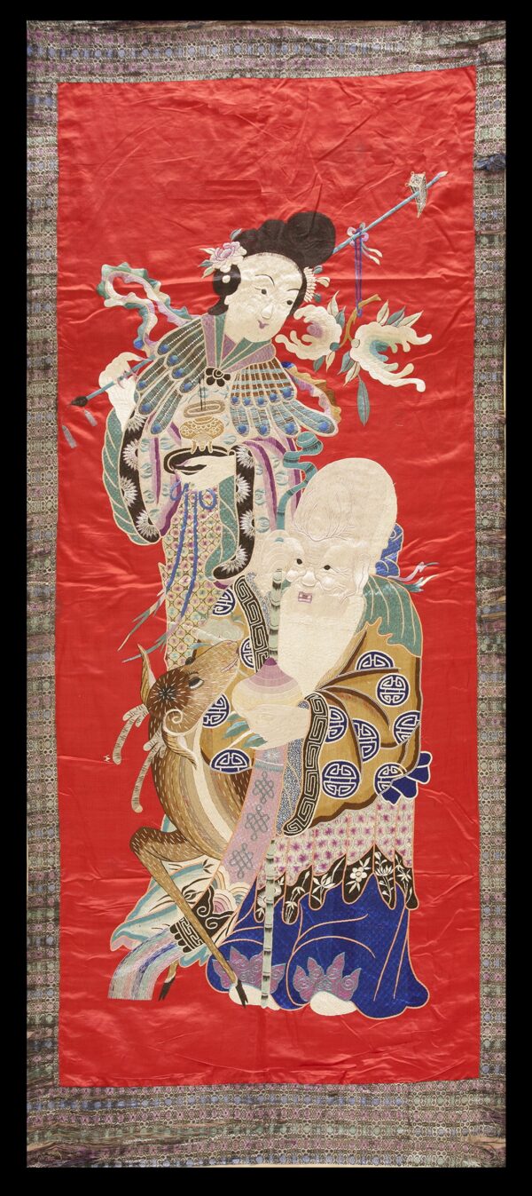 Detailed embroidered red textile with two figures and an animal bordered by a subtle multi-color geometric pattern.