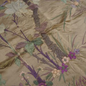 Lustrous tan fabric embroidered with a dense, multicolored botanical pattern of green, purple, and cream elements.