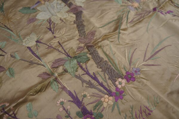Lustrous tan fabric embroidered with a dense, multicolored botanical pattern of green, purple, and cream elements.