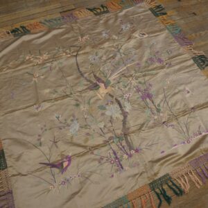 Shiny taupe embroidered textile featuring birds and flowers, surrounded by a colorful fringed edge on wood floors.