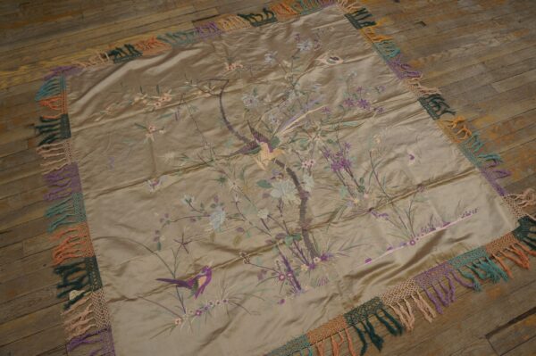 Shiny taupe embroidered textile featuring birds and flowers, surrounded by a colorful fringed edge on wood floors.