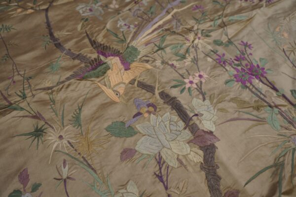 Close-up of a shiny gold textile with embroidered birds, flowers, and foliage in purple, green, and peach.
