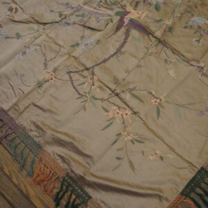 Glossy taupe textile with embroidered flowers and birds, featuring a multicolored tasseled lace edge on wood flooring.
