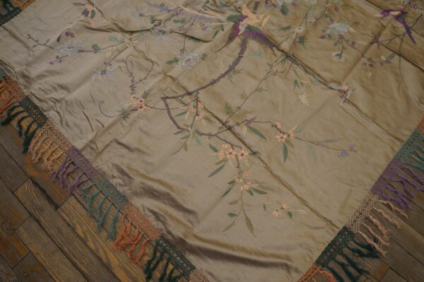 Glossy taupe textile with embroidered flowers and birds, featuring a multicolored tasseled lace edge on wood flooring.
