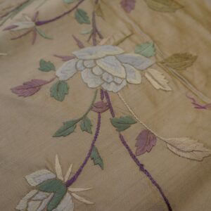 Close-up of a tan textile with embroidered light blue, green, and purple botanical patterns.