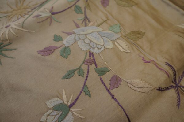 Close-up of a tan textile with embroidered light blue, green, and purple botanical patterns.