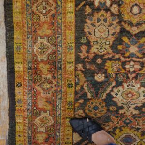 A foot in a black sandal steps on an ornate rug with yellow, red, and dark blue patterns on gray wood floors.