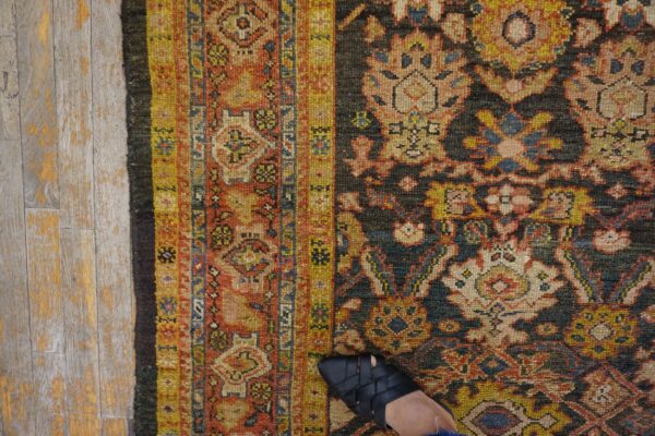 A foot in a black sandal steps on an ornate rug with yellow, red, and dark blue patterns on gray wood floors.
