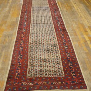 Long traditional runner rug featuring a dense geometric pattern in red, blue, and cream on wood plank flooring.