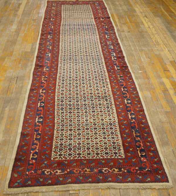 Long traditional runner rug featuring a dense geometric pattern in red, blue, and cream on wood plank flooring.