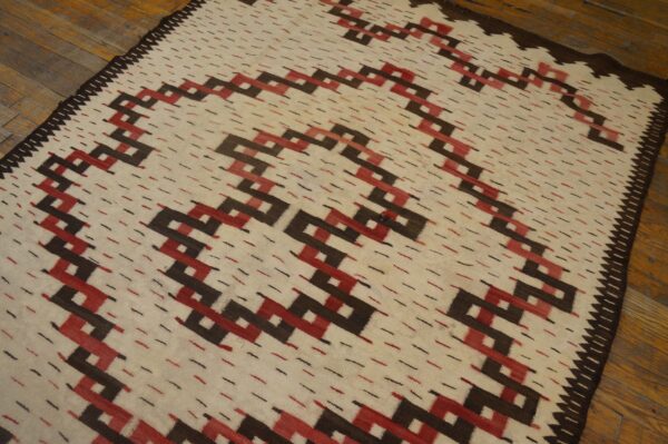 Cream flatweave rug featuring a repeating geometric zig-zag pattern in dark brown and red on a wood floor.