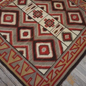 Geometric flatweave rug in red, cream, brown, and grey with a diamond center and arrow motifs on worn wood floor.