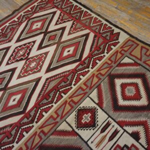 Geometric flat rug with red, cream, brown, and gray diamond patterns on worn wood flooring.