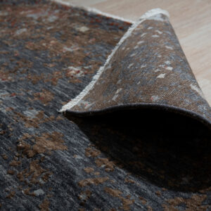 Textured dark brown and gray abstract rug pile detail with white fringe on light wooden planks.