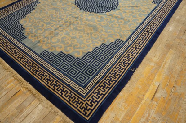 Light blue rug with tan swirling patterns and navy blue greek key borders on worn yellow wood flooring.