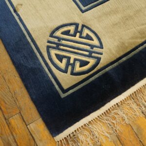 Beige and navy low-pile rug featuring a geometric medallion and fringe over distressed hardwood.