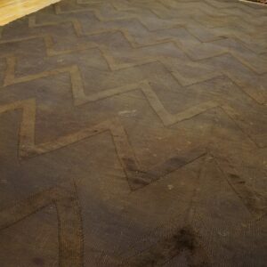 Dark brown textured rug with a repeating zigzag pattern resting on light wooden plank flooring.