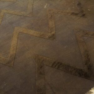 A dark brown rug features a wide chevron pattern in lighter brown textures on visible wood flooring.