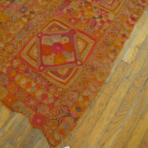 Textured rug displaying complex orange, pink, and gold geometric patterns on worn, light-toned hardwood floors.