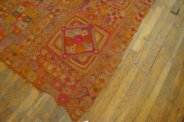 Textured rug displaying complex orange, pink, and gold geometric patterns on worn, light-toned hardwood floors.