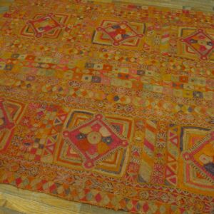 Intricate, embroidered textile featuring geometric patterns in orange, yellow, magenta, and purple laid on wood floors.