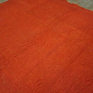 A rust-orange quilted textile rug with golden stitched floral patterns covers rustic wooden flooring.