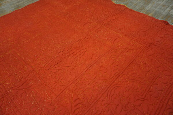A rust-orange quilted textile rug with golden stitched floral patterns covers rustic wooden flooring.