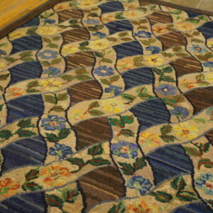 Rustic rug displaying a wavy pattern of beige florals, blue sections, and striped brown geometric shapes on a wood floor.