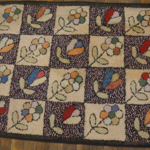 Beige and navy checkerboard hooked rug displaying primitive flowers and leaves on rustic wooden planks.