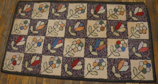 Beige and navy checkerboard hooked rug displaying primitive flowers and leaves on rustic wooden planks.