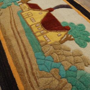 Close-up of a sculpted rug depicting a yellow house and green landscape, bordered in black and orange on wood flooring.